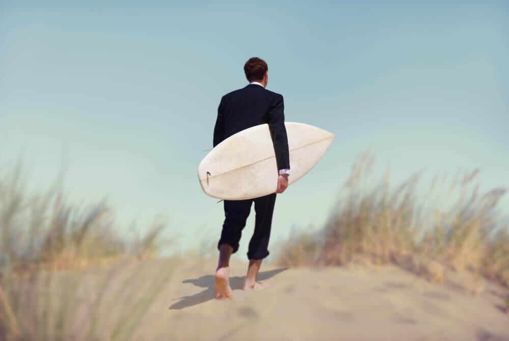 man in suit walking with a surfboard reflecting new profile of the digital nomads
