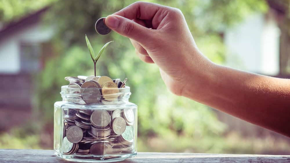 hand dropping a coin into a glass jar full of coins and a seed growing reflecting the need to maximise your hotel marketing budget return