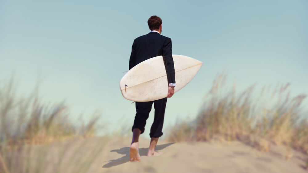 man in a suit with a surfboard on the beach reflecting continuing impact of bleisure travel