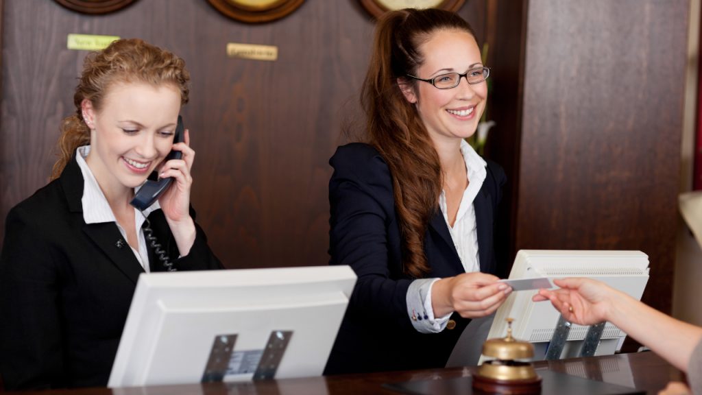 two receptionists at hotel front office with one guest checking in and another on the phone illustrating the potential impact on hotel revenue of front desk upselling